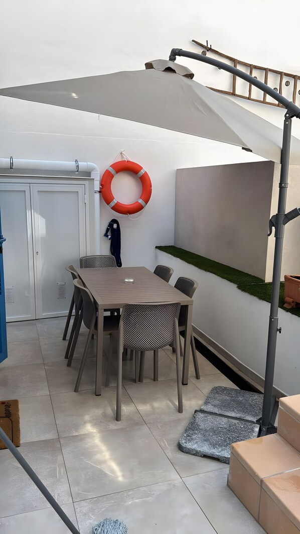 Un espace extérieur abrité avec une table et des chaises, un gilet de sauvetage accroché au mur et une petite étagère pour plantes vertes.