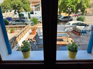 Une fenêtre ornée de deux plantes en pot sur le rebord, une vue sur une rue bordée de voitures garées et, au fond, un immeuble doté d’un balcon.