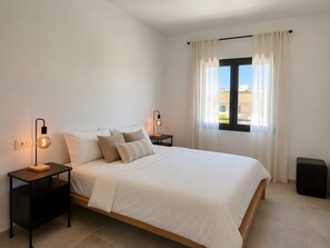 A bedroom with a bed, two bedside tables, a lamp, a window with curtains, and a view of buildings outside.