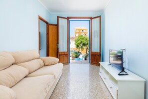 A room with a beige sofa, a television on a white cabinet, and a view of a building and trees through an open door.