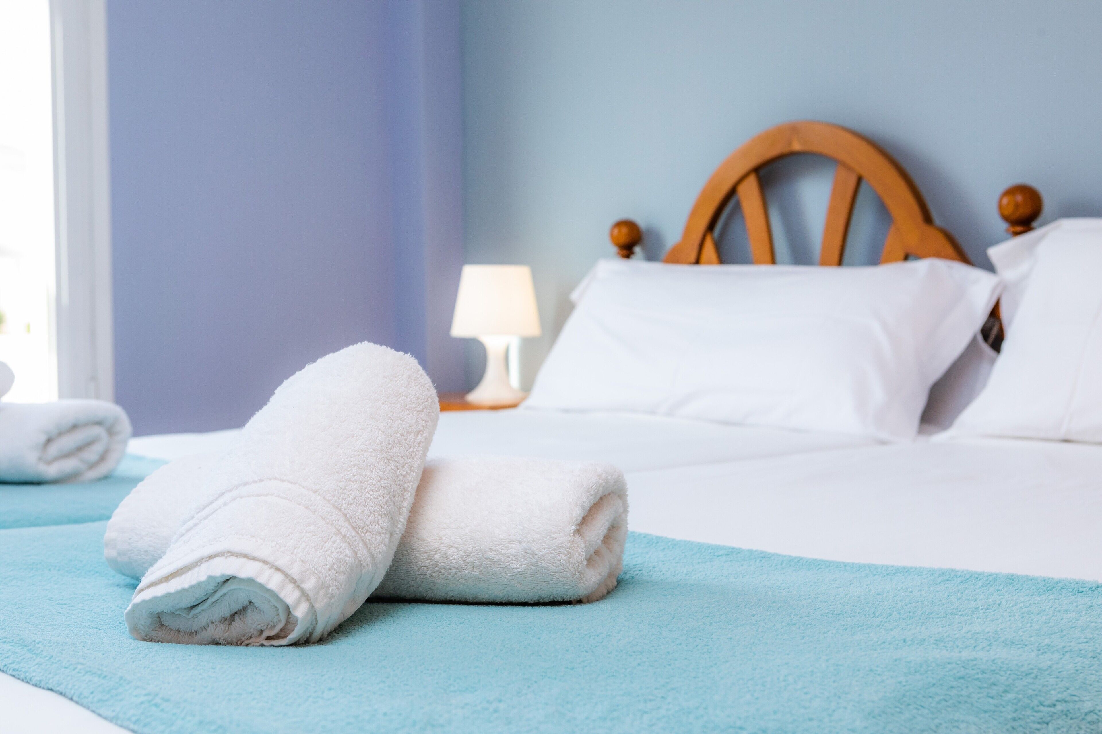 A neatly made bed with white linens and a blue towel, a wooden headboard, and a bedside lamp.