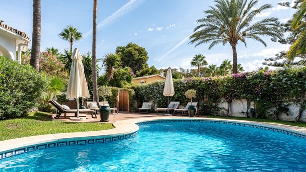 Ein Swimmingpool mit Liegestühlen und Sonnenschirmen, umgeben von Palmen und einem Garten.