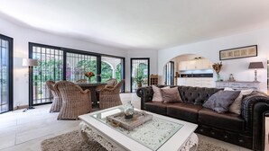 Ein geräumiges Wohnzimmer mit einem Glastisch, einem braunen Sofa, Korbstühlen und Blick ins Freie durch große Fenster.