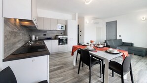 A modern kitchen with a microwave, oven, and sink, a dining area with a table and chairs, and a grey sofa in a living area. - New Apartment Casa Andrea with Lake View, Terrace and On-Site Free Parking (TREZZONE)