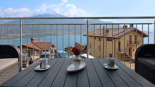 A balcony with a table set for two, overlooking a lake and buildings. - New Apartment Casa Andrea with Lake View, Terrace and On-Site Free Parking (TREZZONE)