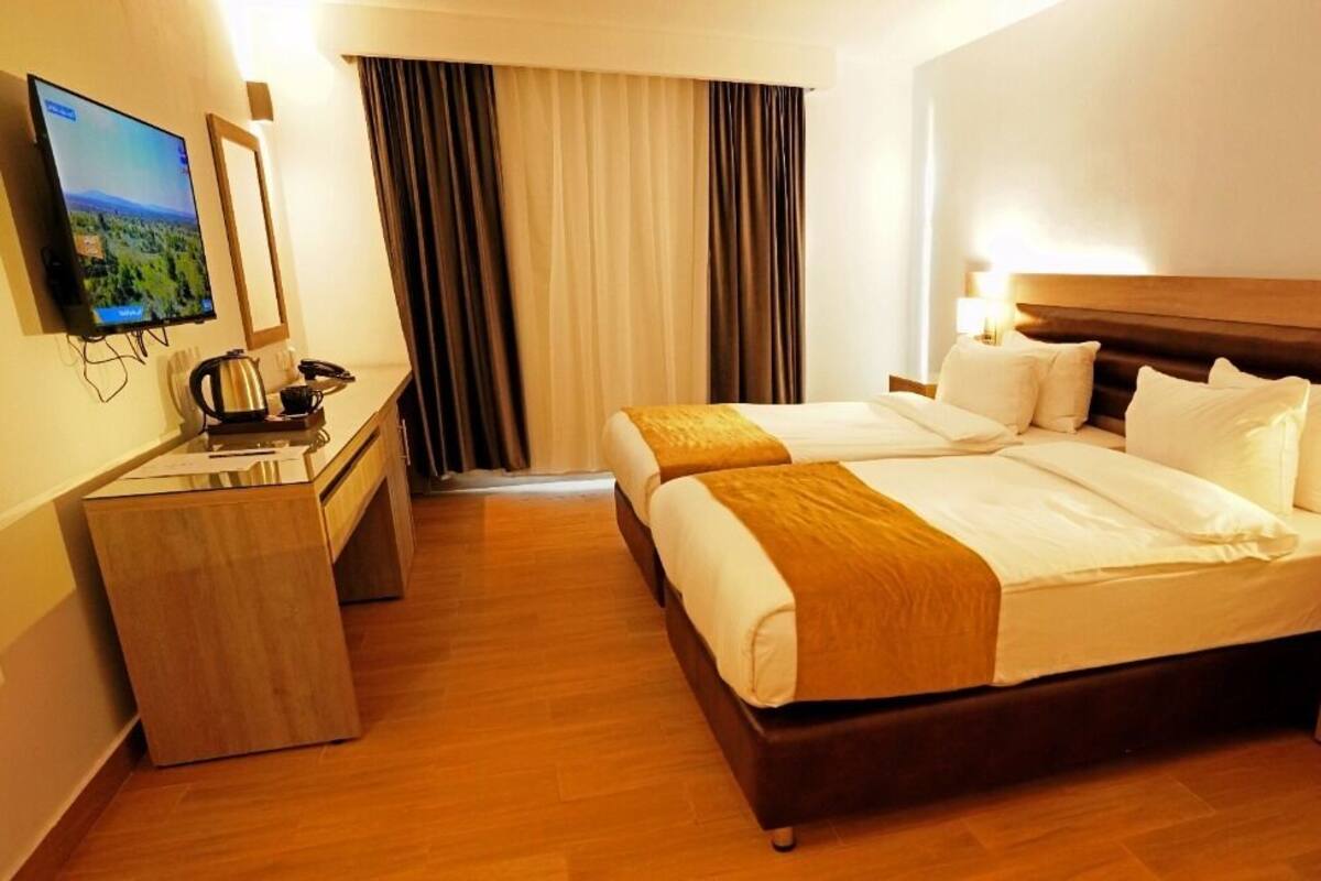 A hotel room with two beds, a desk with a kettle, a TV, and a nightstand.