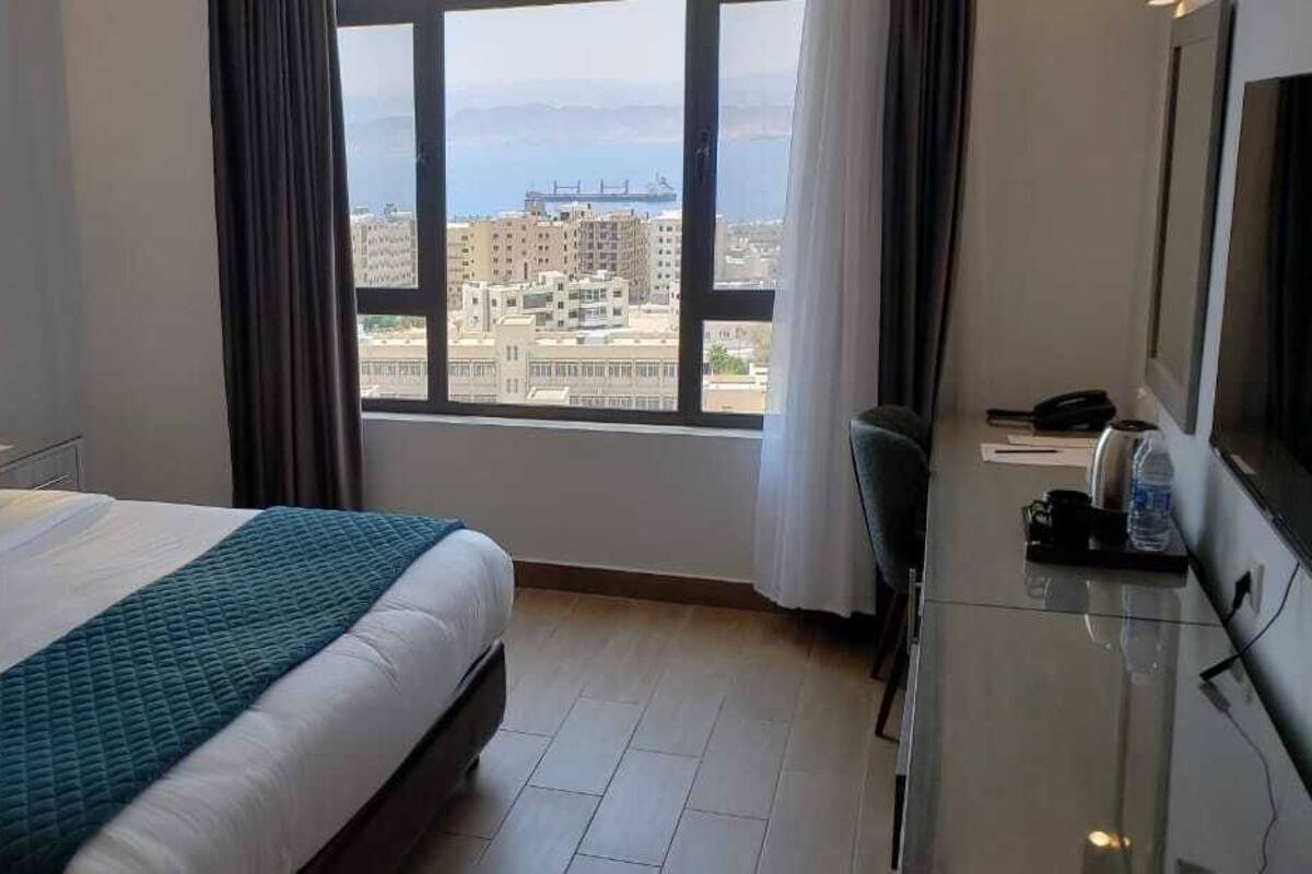 A hotel room with a large bed, a desk with a chair, a TV, and a view of the city through the window.