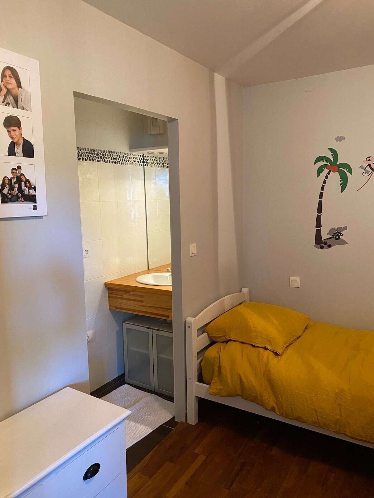 A small bedroom with a bed, a nightstand, a dresser, and a bathroom visible through an open door.