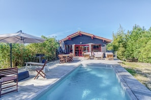 Ein Garten mit Schwimmbecken, Holzterrasse, Gartenmöbeln und einem Haus mit roter Tür.