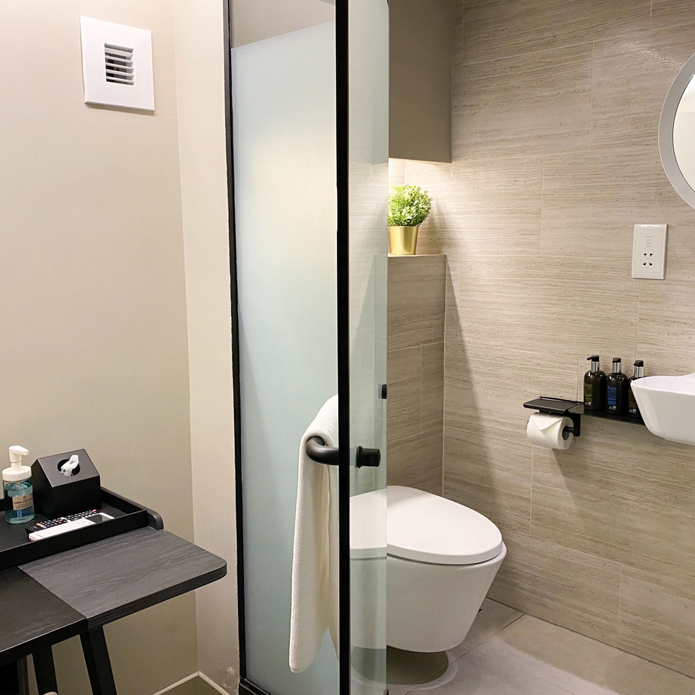 A modern bathroom with a glass shower enclosure, a toilet, a sink with toiletries, and a small table with a towel.