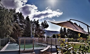 En swimmingpool med ett trädäck runt om, omgiven av ett staket i metall, och ett stort parasoll som ger skugga.