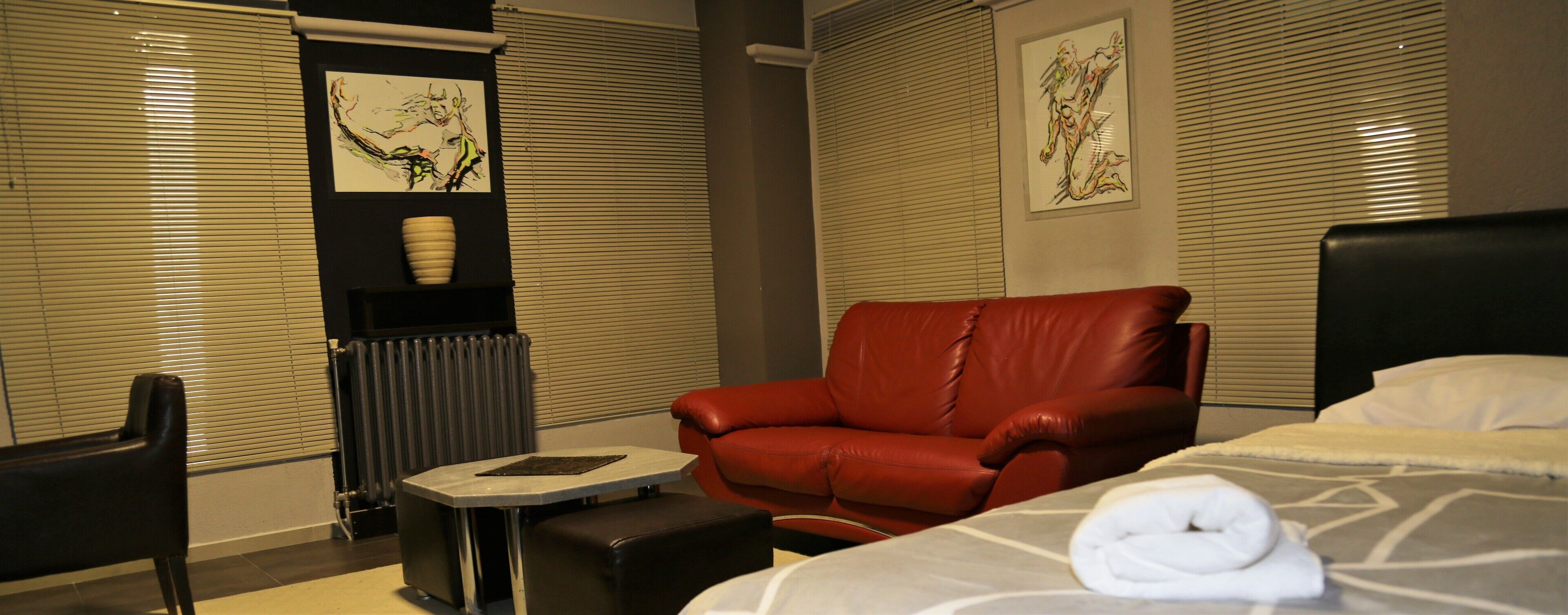Une chambre d’hôtel avec un lit, un fauteuil en cuir rouge, une petite table, un radiateur et des œuvres d’art accrochées aux murs.