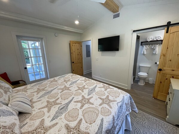 A bedroom with a bed, a TV, a wooden door, and a bathroom with a toilet and towel rack.
