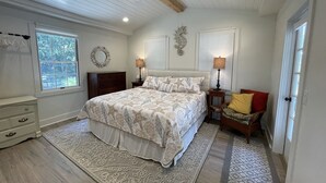 A bedroom with a large bed, two bedside tables with lamps, a chair, a dresser, and a window with blinds.