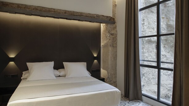 A modern bedroom with a large bed, a stone accent wall, and a large window with curtains. - Mon Suites Fetén (Valencia)