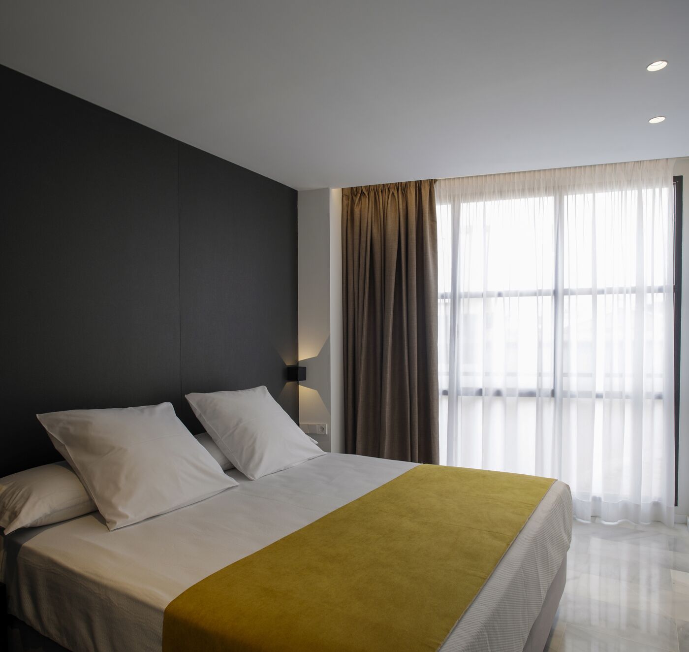 Una habitación de hotel moderna con una cama grande, una pared de acento oscura y un ventanal con cortinas translúcidas.