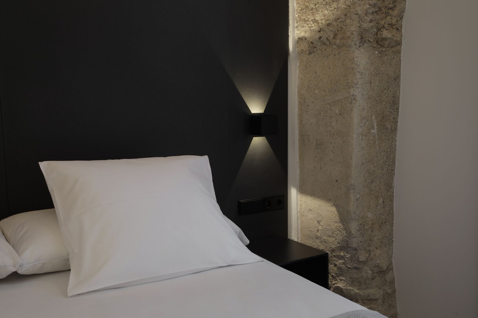 A bed with white bedding and a black nightstand with a wall-mounted light.