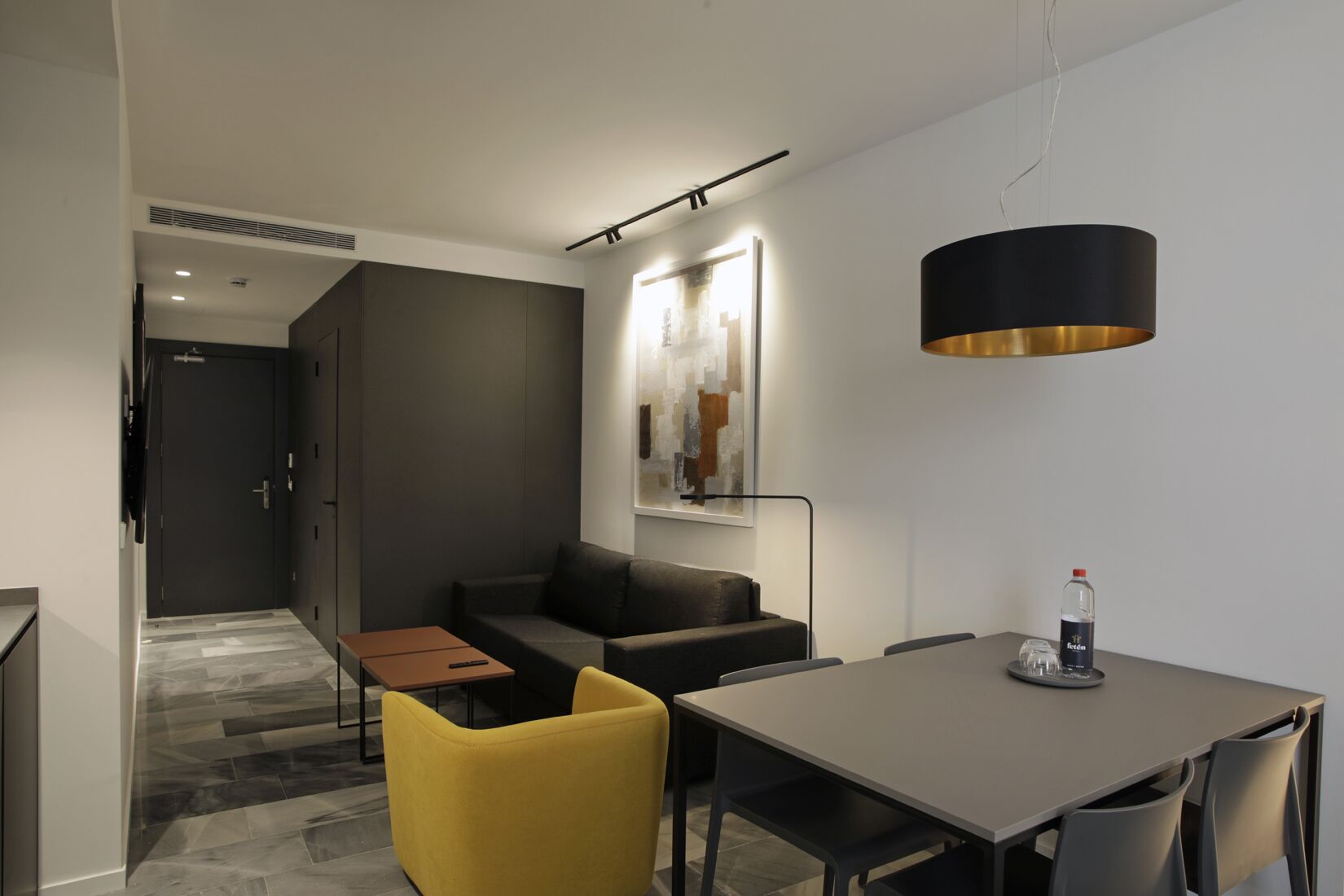 Una habitación de hotel moderna con sofá, una mesa de comedor, una silla amarilla, una lámpara colgante negra y una botella de agua sobre la mesa.