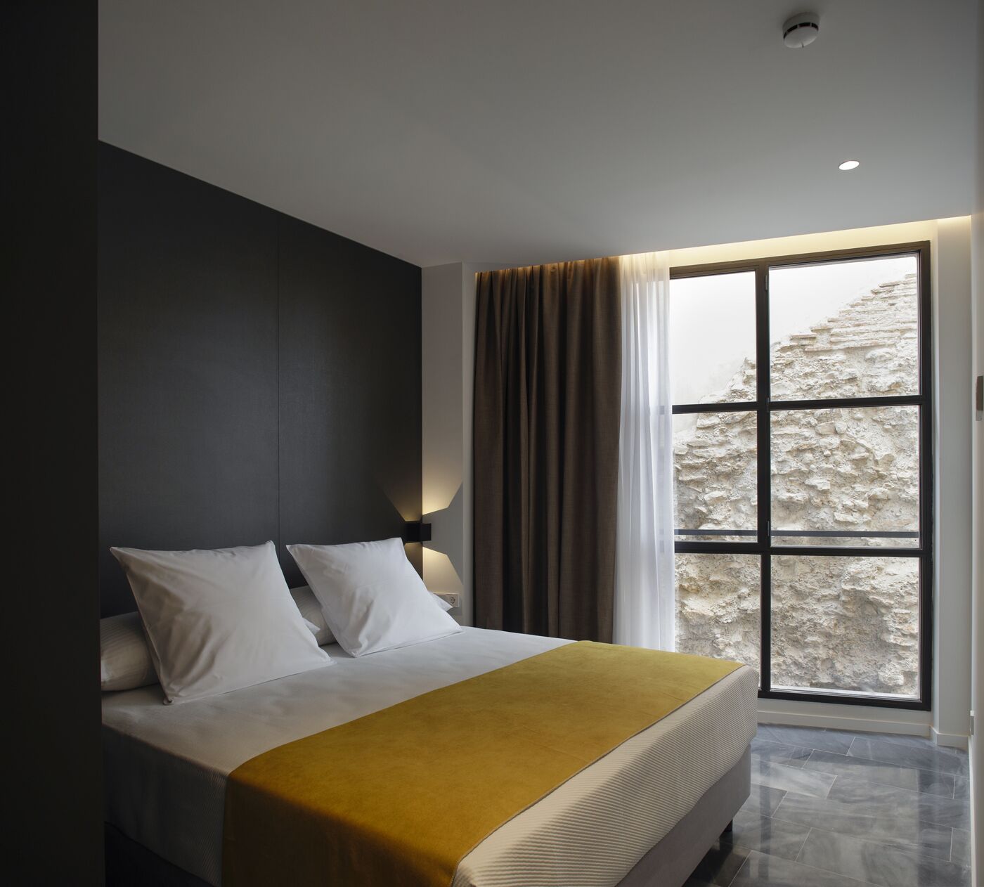 Una camera d'albergo moderna con un letto grande, una finestra con tende e vista su un paesaggio roccioso.