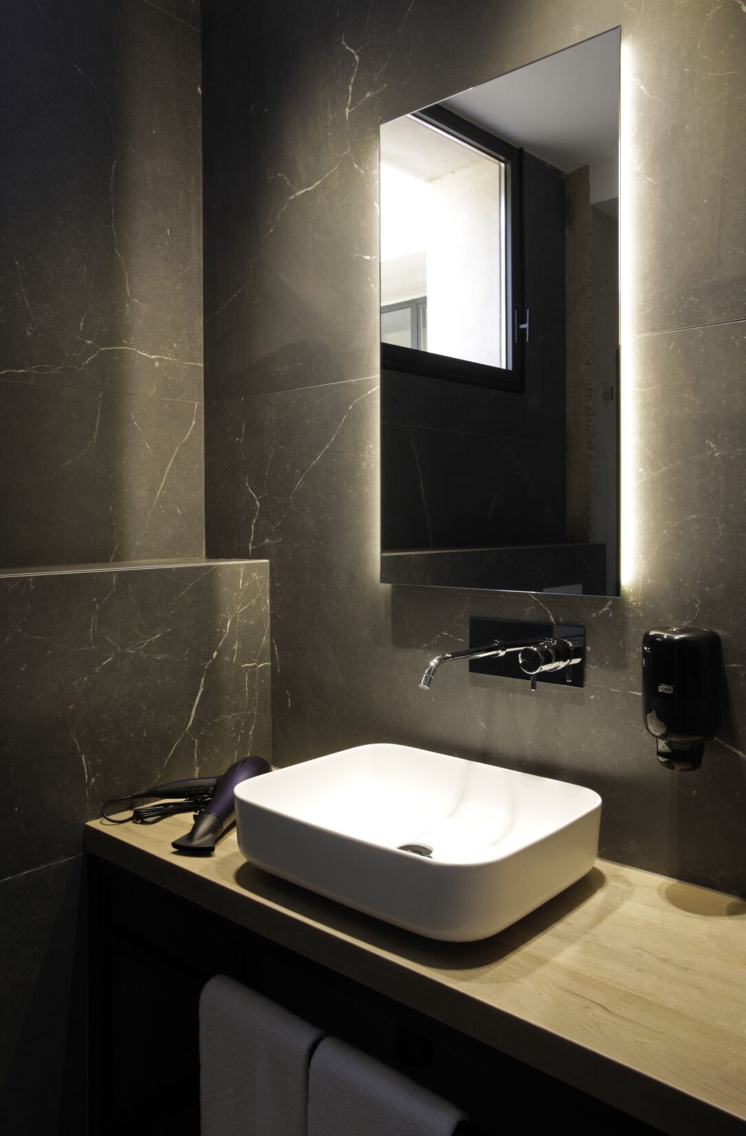 A modern bathroom with a large mirror, a white sink, and a handheld hairdryer on the counter.