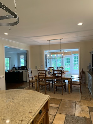 A spacious kitchen with a granite countertop, wooden dining table, and chairs. There is a chandelier hanging above the table, and a large window providing natural light.