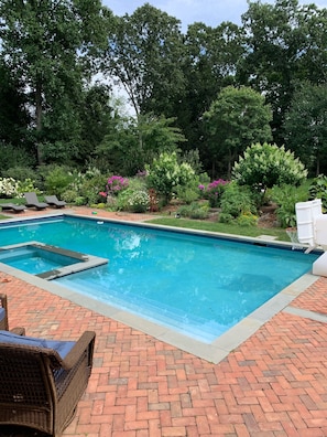 A swimming pool with a brick patio, surrounded by lush greenery and a variety of plants.