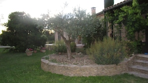 Un jardín con un muro de piedra, diversas plantas y un edificio con techo de tejas.