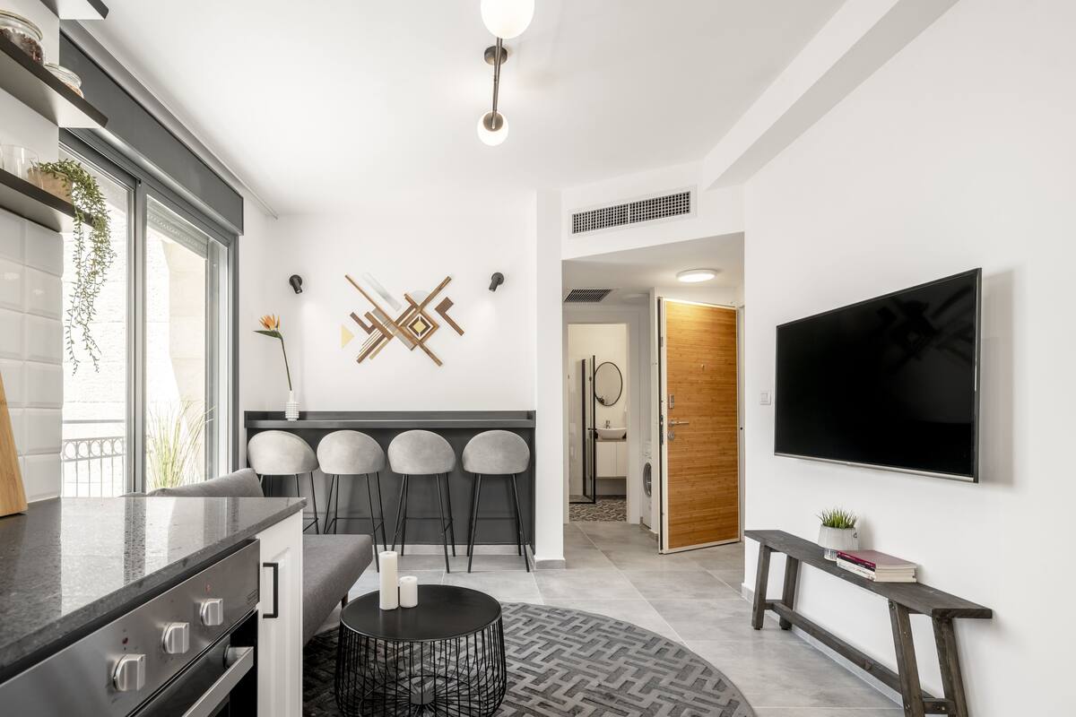 A modern kitchen with a built-in oven, a dining area with three chairs, a round coffee table, and a TV mounted on the wall.