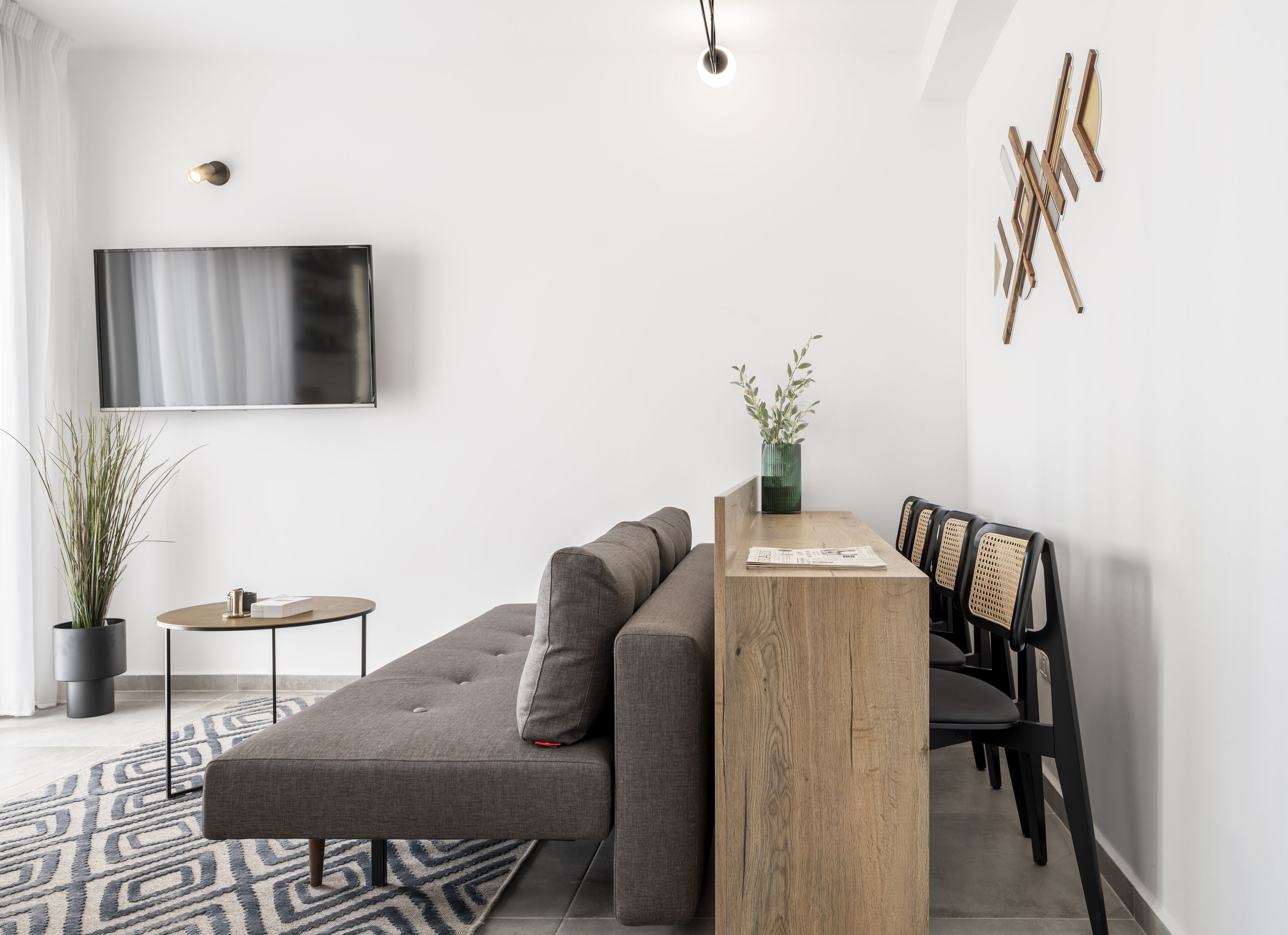 A modern living room with a grey sectional sofa, a wooden coffee table, a TV mounted on the wall, and a dining area with a wooden table and chairs.