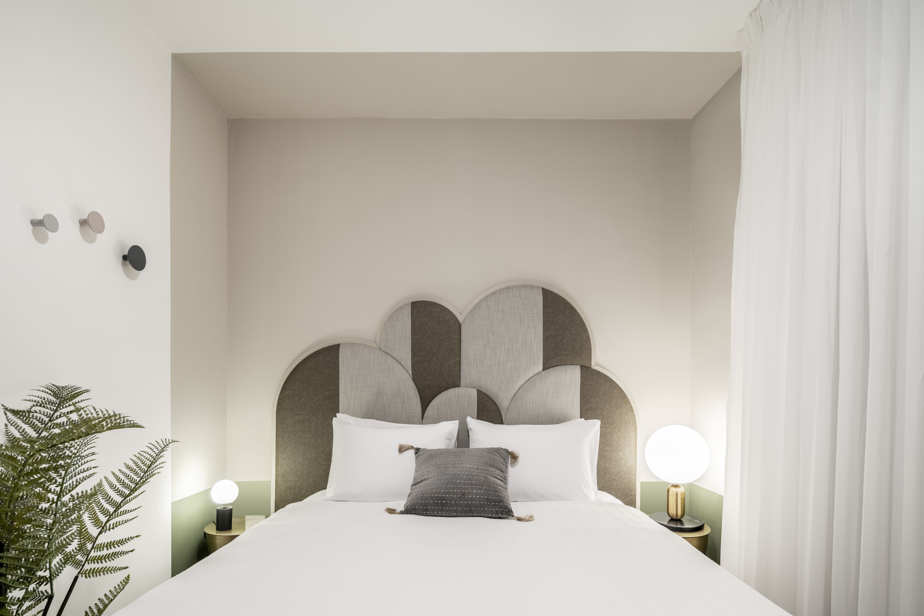 Una cama bien tendida con sábanas blancas y un almohadón gris, un cabecero moderno con diseños geométricos y una pequeña planta en maceta a la izquierda.
