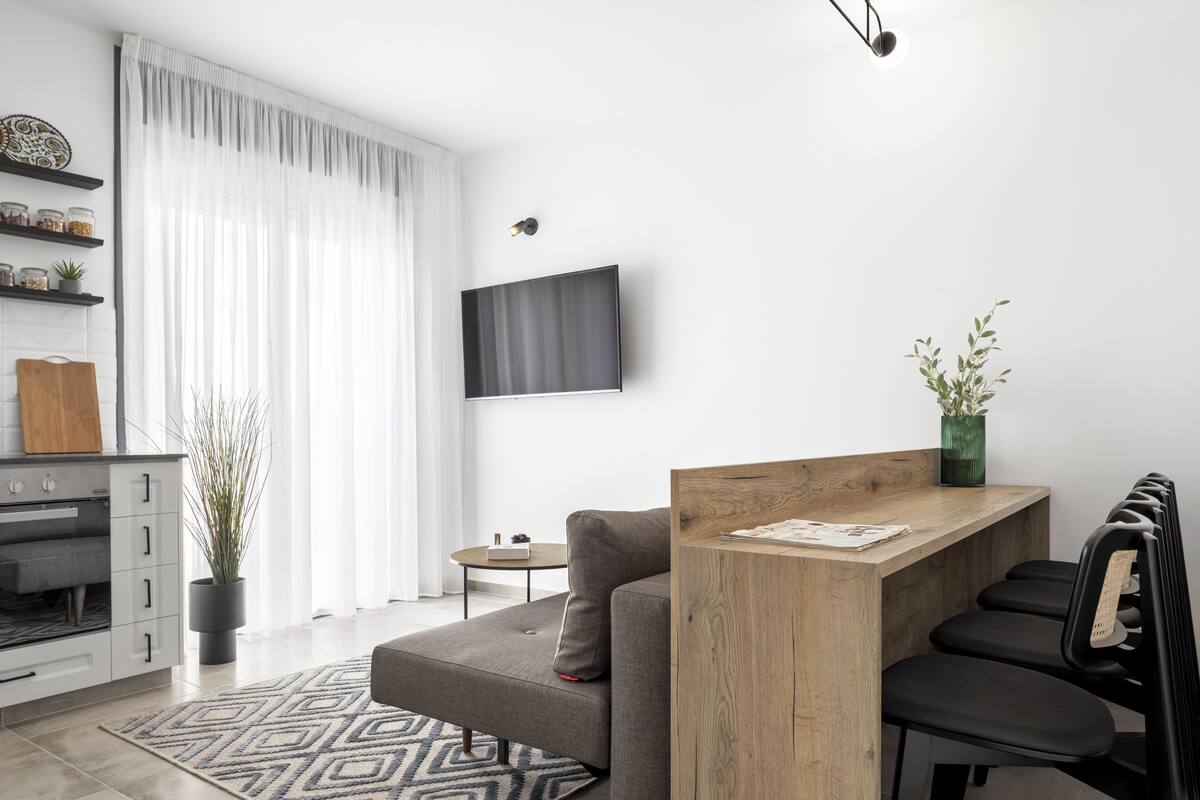 A modern living room with a grey sofa, a wooden coffee table, a flat-screen TV mounted on the wall, and a potted plant.