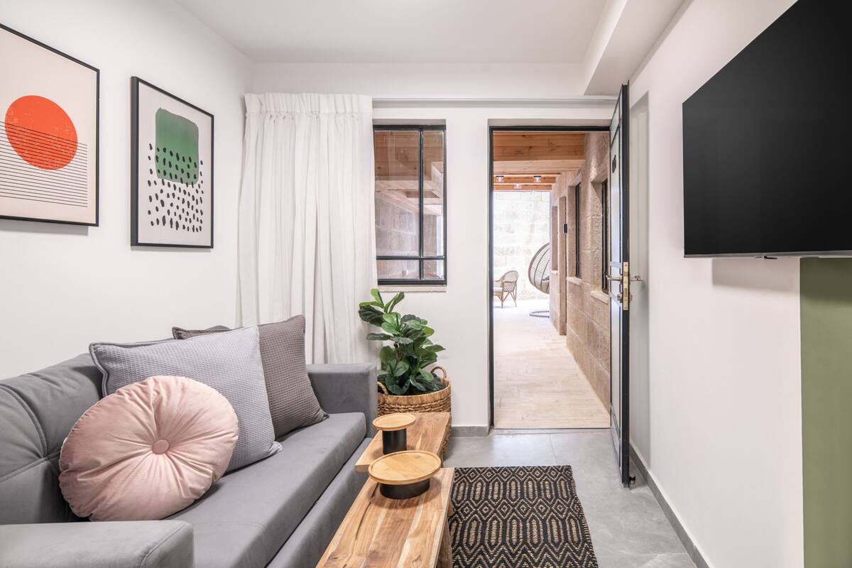 A modern living room with a grey sofa, a wooden coffee table, and a large TV. There are two framed artworks on the wall, a potted plant, and a window with white curtains.