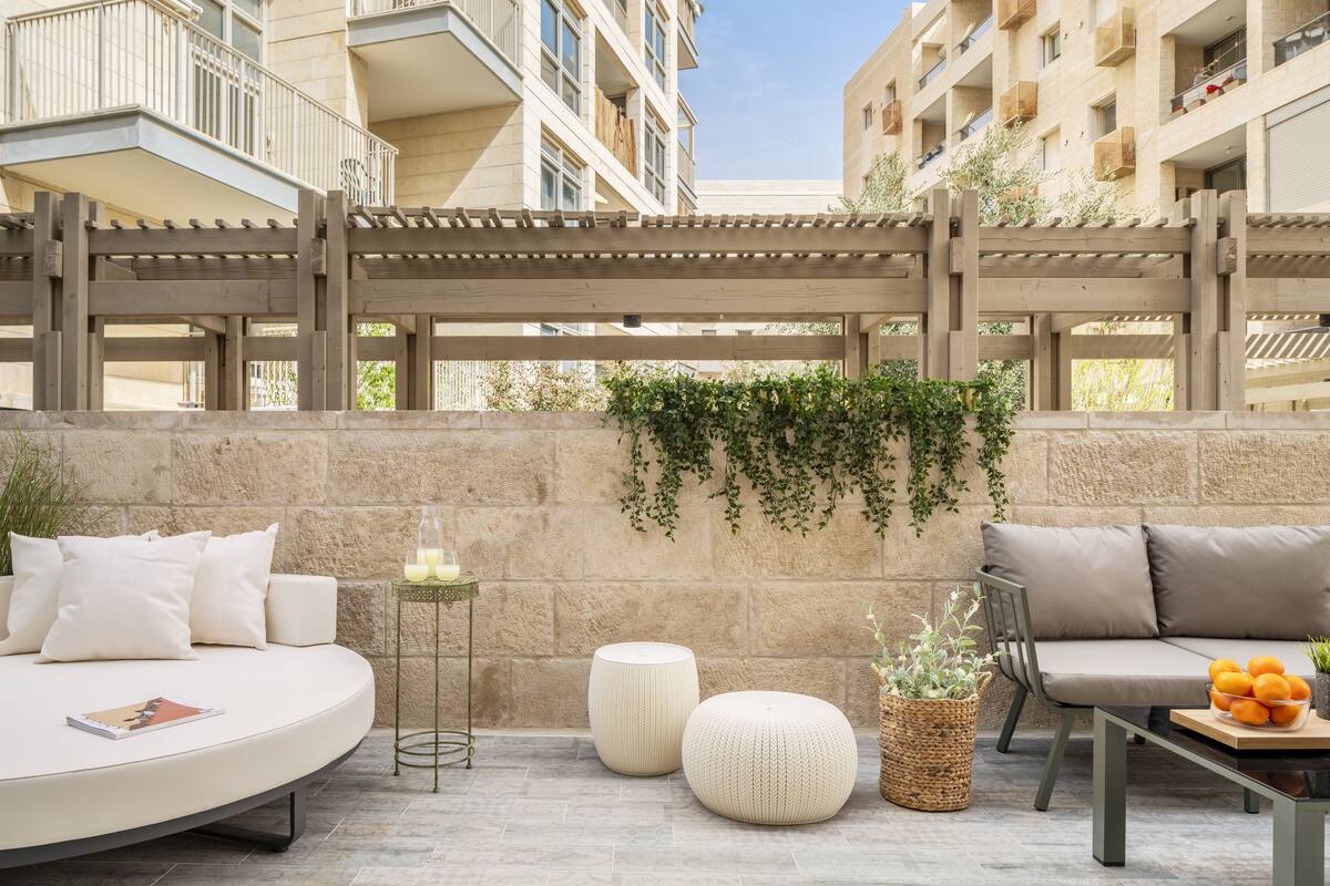 A modern outdoor seating area with a round white sofa, a wooden pergola, and a stone wall with greenery.