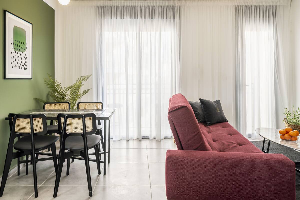 A modern living room with a red sofa, a dining table with chairs, and a large window with sheer curtains.