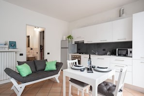 A modern kitchen with white cabinets, a dining table with chairs, and a grey sofa with green cushions.