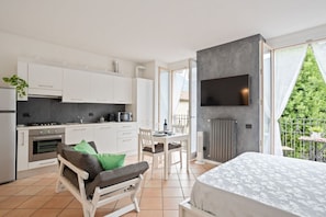 Une cuisine moderne avec des meubles blancs, un coin repas comprenant une table et des chaises, un lit recouvert d’une couvre-lit à motifs et un téléviseur fixé à un mur gris.
