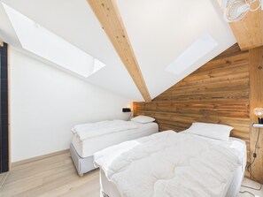 Une chambre avec deux lits, des poutres apparentes en bois au plafond, un puits de lumière et un luminaire fixé au mur.