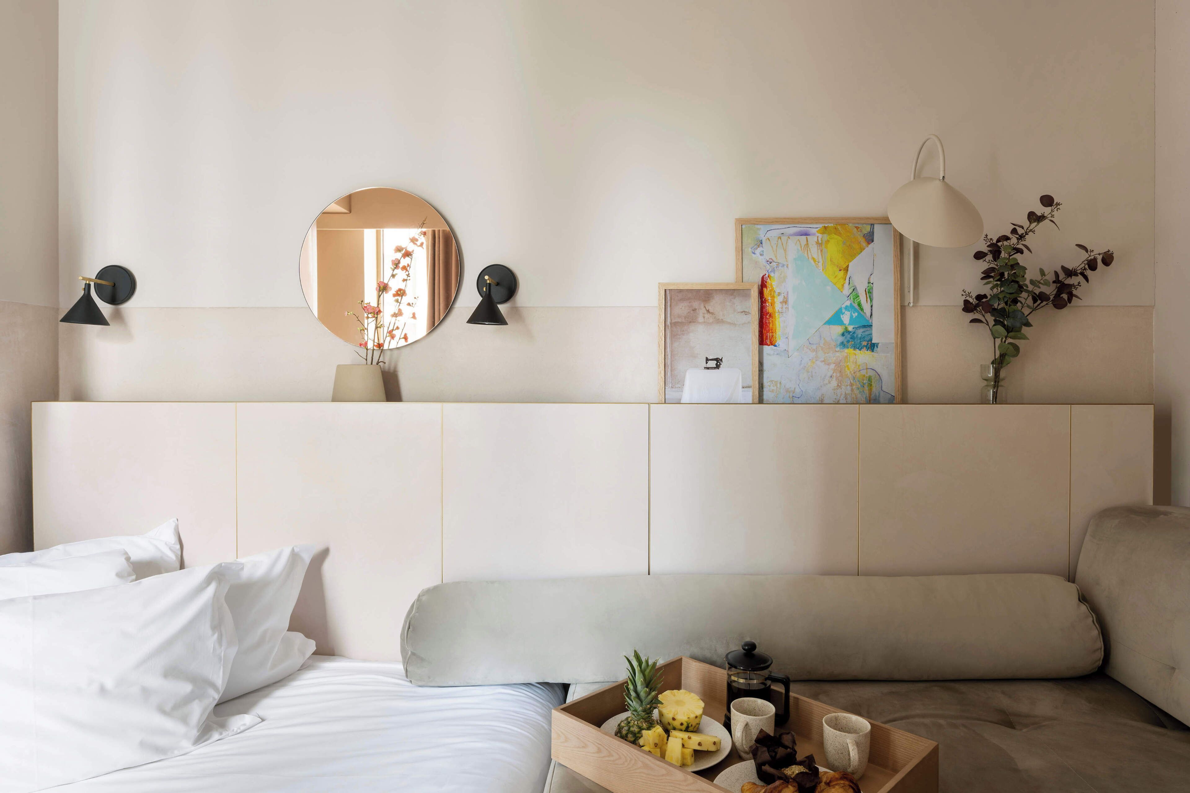 A hotel room with a bed, a bedside table with a tray of fruit and a cup, a round mirror, and wall-mounted lamps.