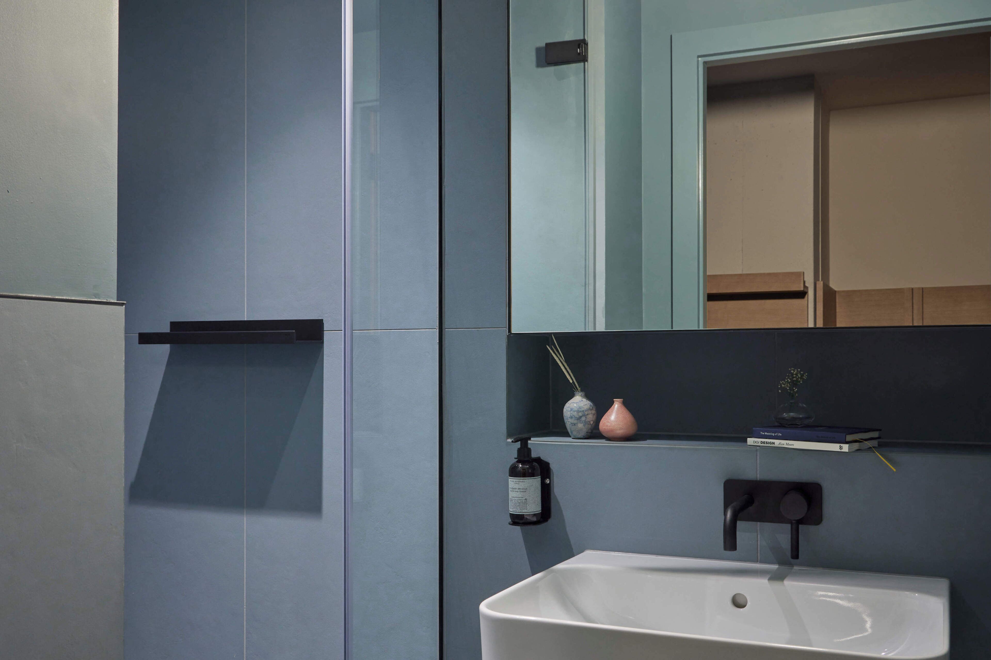 A bathroom with a white sink, dark wall tiles, a mirror, and a shelf with decorative items.