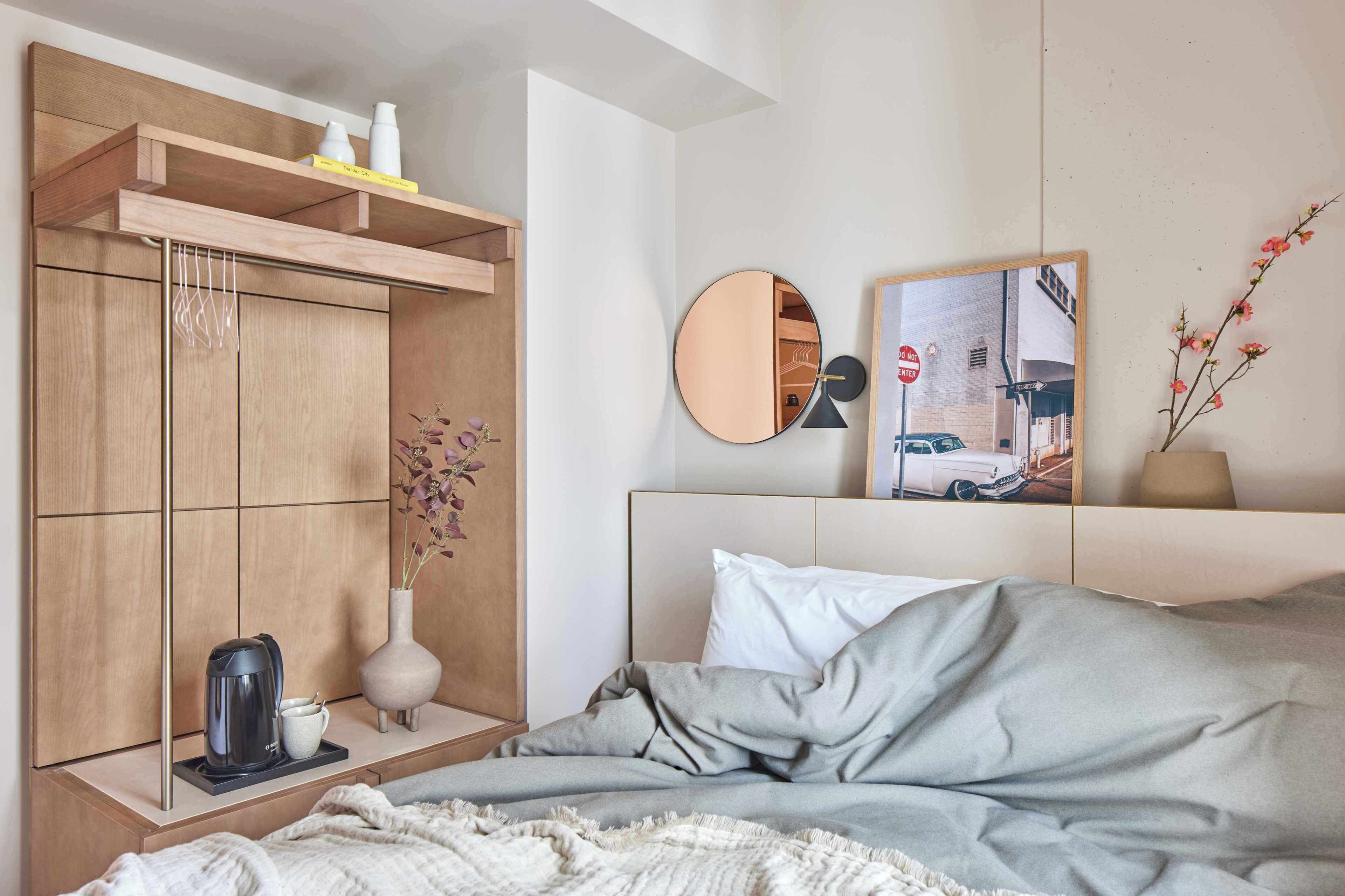 Ein Schlafzimmer mit einem Bett, einem Nachttisch mit Kaffeemaschine, einem Spiegel und einem an der Wand befestigten Regal mit Büchern und Flaschen.