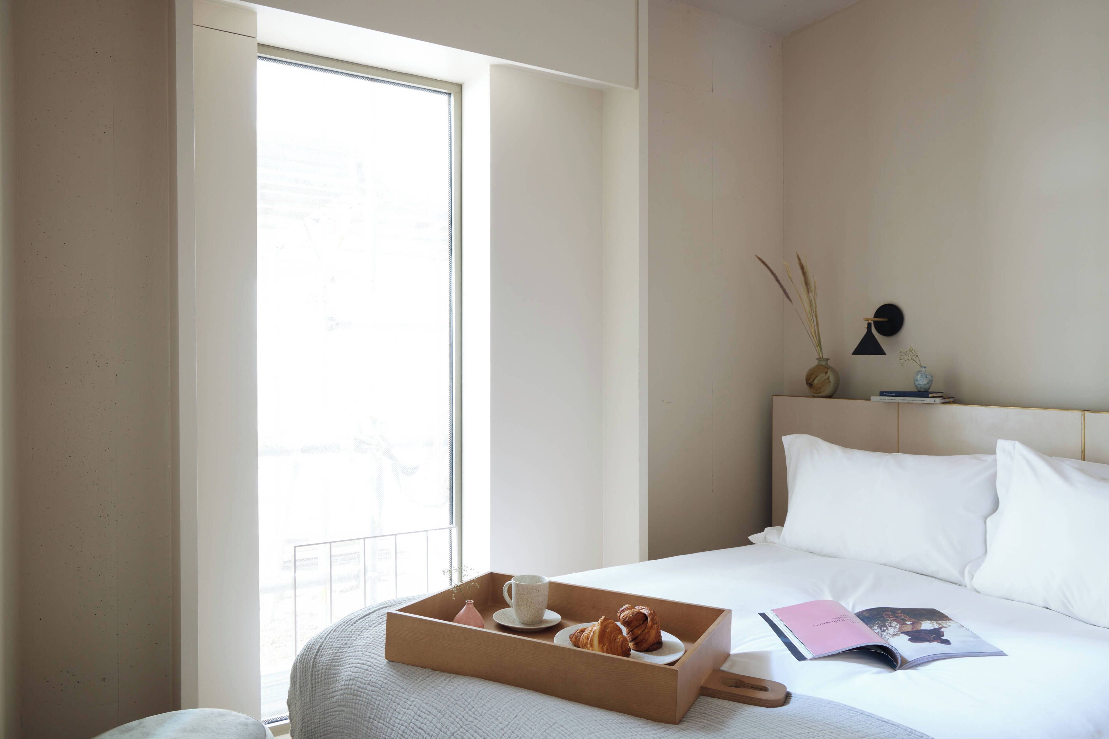 A neatly made bed with pillows, a bedside table with a lamp, a window with a view, and a tray with pastries.