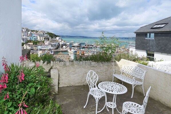 A balcony with white metal chairs and a table, overlooking a coastal town with buildings and boats.