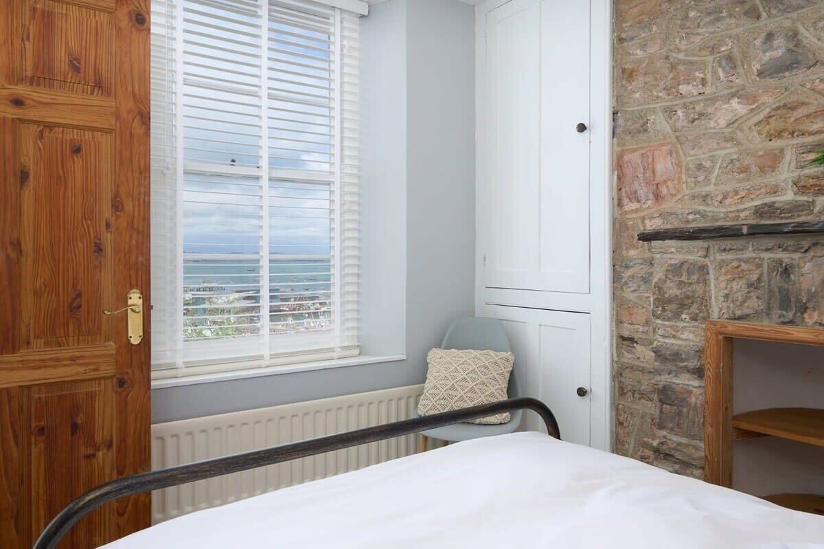 A bedroom with a wooden door, a window with blinds, a radiator, a bed, a chair, and a stone wall.