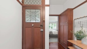 Een houten deur met een geometrisch glasraam, een bankje met een bloempot en een blik op een slaapkamer met een bed en raam.