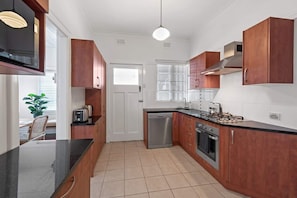 A modern kitchen with wooden cabinets, a black countertop, stainless steel appliances, and a window with blinds. - Art Deco in Ascot! ~ 2 Bed / 1 Bath / 1 Car (Ascot)