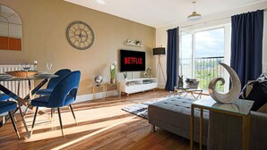 A modern living room with a flat-screen TV, a dining area with blue chairs, a sofa, and a large window with curtains. - Luxury Apartment with Balcony, Parking & Fast Wifi (Milton Keynes)