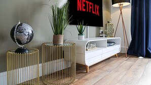 A modern living room with a white entertainment unit, a globe on a gold stand, a potted plant, and a large Netflix sign on the wall. - Luxury Apartment with Balcony, Parking & Fast Wifi (Milton Keynes)