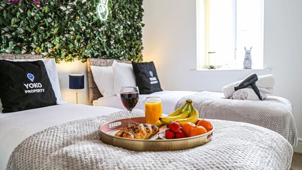 A hotel room with two beds, a breakfast tray with croissants, fruit, and juice, and a wall decorated with greenery and a logo. - Luxury Apartment with Balcony, Parking & Fast Wifi (Milton Keynes)