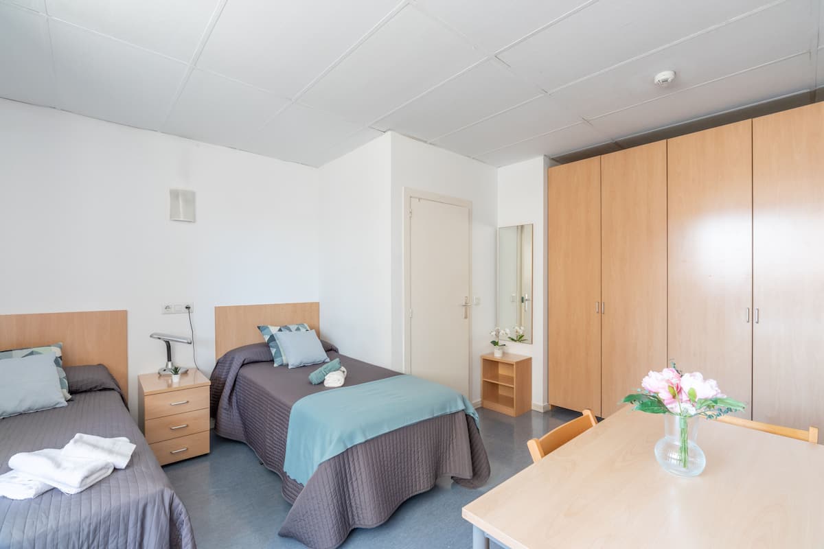 Hotellihuone, jossa on kaksi erillistä sänkyä, puinen vaatekaappi, pieni pöytä maljakko täynnä kukkia ja työpöytä lampun kanssa.