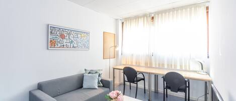 A modern living room with a grey sofa, a wooden coffee table, and a desk with two chairs. There is a painting on the wall and a window with curtains.